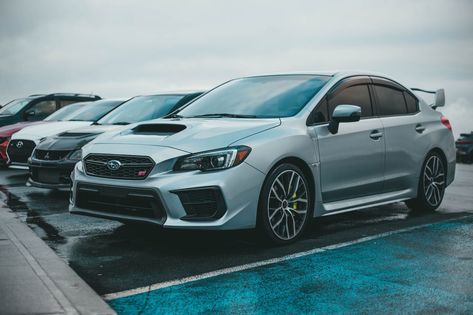 A silver Subaru WRX parked in a city parking lot with other cars on a rainy day.